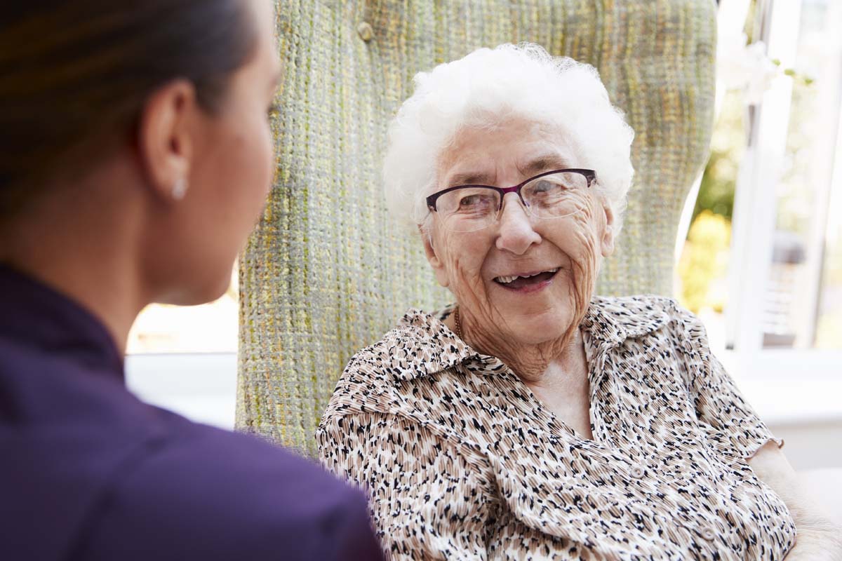Female Resident Sitting In Chair And Talking With Carer In Retirement Home